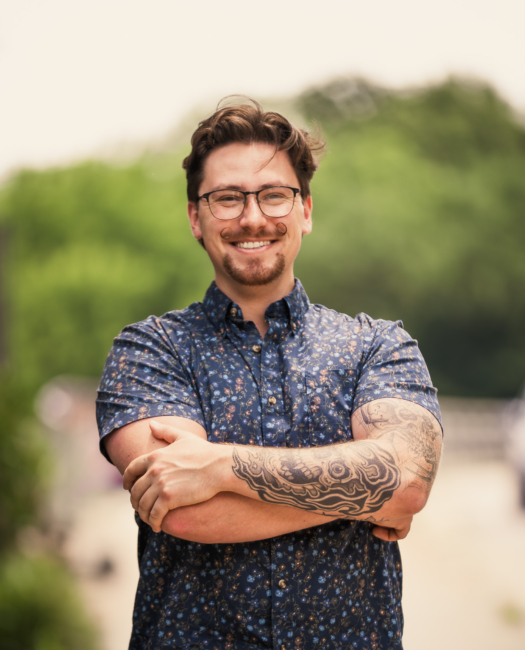 Smiling Wye Hill Kitchen & Brewing team member with glasses and arm tattoos, arms crossed, outdoors in downtown Raleigh